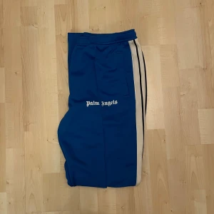 Blå Palm Angels mjukisbyxor med vit stripe - Snygga blå mjukisbyxor från Palm Angels med vit sidostripes och logga tryckt på framsidan. Byxorna har elastisk midja och raka ben, perfekt för en chill streetwear-look. Materialet är mjukt och skönt, troligtvis polyester. Klassisk design med fickor på sidorna.