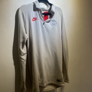 Vit långärmad PSG fotbollströja Nike - Snygg vit långärmad fotbollströja från Nike med PSG-logga på bröstet och röd Nike-swoosh. Tröjan har halv dragkedja och detaljer i rött vid kragen. Perfekt för dig som gillar Paris Saint-Germain och vill ha en stilren look.