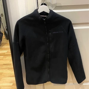 Svart fleecejacka från Röhnisch  - Svart fleecejacka från Röhnisch i storlek XS. Jackan har hel dragkedja framtill, hög krage och en bröstficka med dragkedja. Broderad logga i lila på bröstet. Perfekt för lager-på-lager och chill dagar.