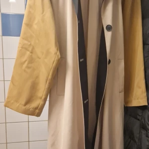 Beige trenchcoat från ONLY med kontrastkrage - Snygg beige trenchcoat från ONLY med mörkblå krage och knappar. Jackan har långa ärmar i en ljusbrun ton och ett svart bälte i midjan. Klassisk design med två fickor framtill och markerad krage som ger en cool kontrast.