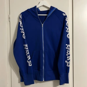 Blå hoodie med tryck och dragkedja - Snygg blå hoodie med vita tryck längs ärmarna och huvan. Tröjan har hel dragkedja framtill, huva och ribbade muddar vid ärmsluten. Perfekt för dig som gillar streetwear och vill sticka ut lite extra. Materialet känns mjukt och bekvämt.