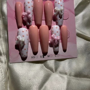 Söta rosa och vita lösnaglar - Set med 10 långa lösnaglar i ballerinaform, hälften i ljusrosa och hälften vita med rosa prickar. Fyra naglar har 3D-rosetter i vitt och rosa.  Perfekt för dig som vill ha gulliga och trendiga naglar. Handgjorda press on nails. Storlek S