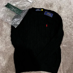 Svart stickad tröja Polo Ralph Lauren - I always send it same day! Never used! Svart stickad tröja från Polo Ralph Lauren med klassisk kabelstickning och röd broderad logga på bröstet. Rund halsringning och långa ärmar. Tröjan har ribbade muddar vid ärmslut och nederkant. Perfekt för dig som gillar stilrena och tidlösa plagg. 