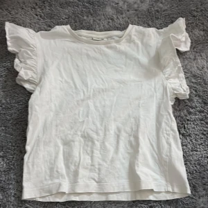 Vit volangärmad topp från Kappahl - Supersöt vit topp från Kappahl i storlek 146/152. Toppen har korta ärmar med volangdetaljer som ger en extra touch. Tillverkad i mjuk bomull och passar perfekt till både jeans och kjol. Enkel och stilren design som funkar till många outfits.