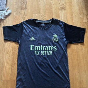 Real Madrid Benzema Adidas fotbollströja - Snygg mörkblå Real Madrid fotbollströja från Adidas med Benzema och nummer 9 på ryggen. Tröjan har korta ärmar, La Liga-märke på ärmen och klubbmärke samt Adidas-logga på bröstet. Tillverkad i lätt och ventilerande material, perfekt för match eller träning.