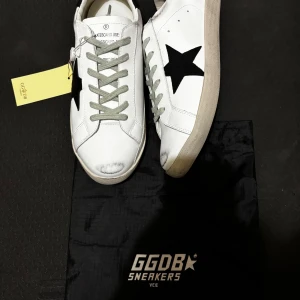 Golden Goose skor - Golden Goose sneakers i vitt skinn med svart stjärna på sidan och svart häl med logotyp. Grå snörning och grå mockadetaljer på hälen. Platt sula och rund tå. Snygg, urban stil med kontrastdetaljer och klassisk siluett.