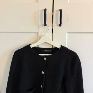 Svart kofta från Gina Tricot - Stilren svart kofta från Gina Tricot med rund halsringning och dekorativa silvriga knappar framtill. Koftan har två fickor och är tillverkad i ett grovstickat material som ger en mysig känsla. Perfekt att slänga på sig över en t-shirt eller topp.