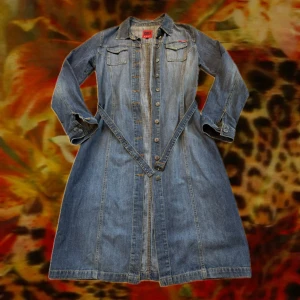 Lång jeansjacka från ONLY - Snygg lång jeansjacka från ONLY med klassisk skjortkrage, långa ärmar och knappar hela vägen framtill. Den har två bröstfickor med lock och ett smalt bälte i midjan för att framhäva siluetten. Perfekt för dig som gillar denim och vill sticka ut.