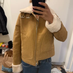 Beige pälsjacka med dragkedja - Snygg beige pälsjacka med mjuk fuskpäls på krage och ärmslut. Jackan har två spännen vid halsen och dragkedja framtill. Perfekt för kalla dagar och ger en cool vintage vibe.