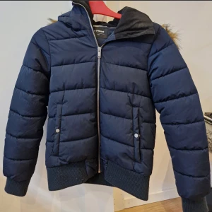 Mörkblå pufferjacka från Everest- barn - Säljer en mörkblå pufferjacka från Everest med huva och fuskpälsdetalj. Jackan har dragkedja framtill, två fickor med tryckknappar och ribbade muddar. Perfekt för kalla dagar och riktigt skön att bära.