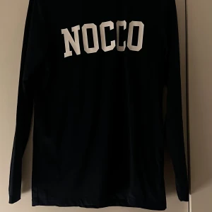 Svart långärmad NOCCO-tröja S - Svart långärmad tröja från NOCCO med stor vit logga på ryggen. Tillverkad i mjuk funktionell och stretchig polyesterblandning som passar perfekt till träning eller chill. Enkel design med rund hals och skön passform.