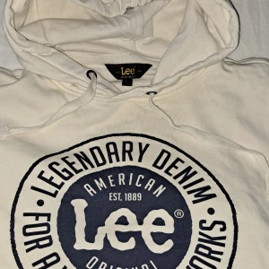 Beige hoodie från Lee med tryck - Säljer en vit hoodie från Lee med stort mörkblått tryck på bröstet. Tröjan har huva med snörning och är i mjuk bomull. Regular fit och perfekt för dig som gillar streetwear vibes.