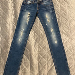 Blå skinny jeans från Nudie Jeans - Säljer ett par blå skinny jeans från Nudie Jeans med slitna detaljer och snygga kontrastsömmar. Modellen har klassisk femficksdesign, orangea sömmar och Nudie-logga på bakfickan. Perfekt passform för dig som gillar smal siluett.