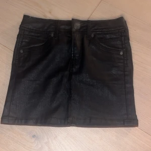 Svart jeanskjol från Pepe Jeans - Svart jeanskjol gjord för barn från Pepe Jeans med klassisk femficksdesign och snygga nitar på bakfickorna. Kjolen har bälteshällor och stängs med knapp och dragkedja. Materialet är en mix av bomull, polyamid och elastan för en bekväm passform. Storlek 116(6)