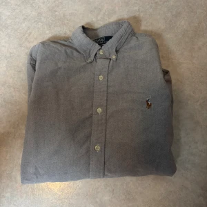 Grå skjorta Polo Ralph Lauren L - Slim fit grå skjorta från Polo Ralph Lauren med klassisk button-down krage och broderad logga på bröstet. Skjortan har långa ärmar, vita knappar och är tillverkad i mjuk bomull. Perfekt för dig som gillar stilren och enkel design.