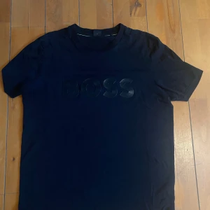 Svart BOSS t-shirt med logga - Svart t-shirt från BOSS med diskret glansig logga framtill. Klassisk rund halsringning och korta ärmar. Snygg och stilren modell som passar perfekt till jeans eller shorts. Materialet känns mjukt och skönt mot huden.