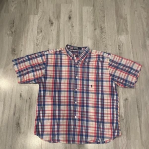 Rutig kortärmad skjorta Polo XL - Snygg rutig skjorta från Polo i storlek XL, med klassiskt mönster i blått, rött, vitt och inslag av beige. Skjortan har korta ärmar, knappar framtill och en bröstficka med Polo-loggan broderad. Tillverkad i 100% bomull för en skön och luftig känsla.