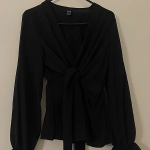 Svart omlottblus med knyt från SHEIN - Svart omlottblus från SHEIN med långa ärmar och snygg knytning framtill. Blusen har v-ringad hals och ballongärmar med resår vid ärmslut. Perfekt för dig som gillar stilrena och trendiga plagg.