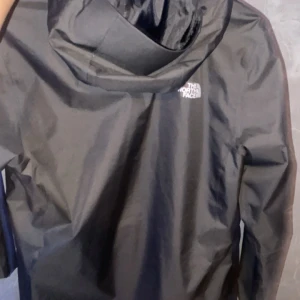 Svart regnjacka från The North Face - Svart regnjacka från The North Face med huva och dragkedja framtill. Jackan har en stilren look med logga på bröstet och ryggen. Perfekt för blöta dagar och har en normal passform med långa ärmar. Materialet är syntet och designen är enkel och funktionell. Storlek M nästan helt ny använd 3-2 gånger. Nästan inga tecken på användning. Kan gå ner i pris vid SNABB affär!