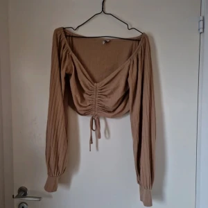 Croppad långärmad topp med snörning - Säljer en beige croppad topp med långa ärmar och snygg snörning framtill från Nelly i storlek M.. Lite fläckar från tvättmedel på baksidan men inget som syns super tydligt och det skulle säkert gå bort om man tvättar om den. Katt finns i hemmet