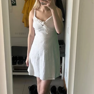 Vit sommarklänning från Hollister - Superfin vit klänning från Hollister med broderade blommor, knyt framtill och smala axelband. Klänningen är kort och har en lätt utsvängd passform, perfekt för varma dagar. Tillverkad i bomull för en luftig känsla.