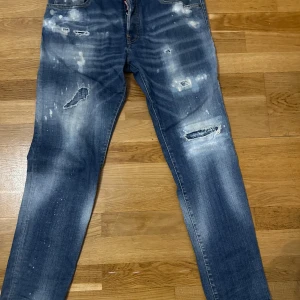 Dsquared2 blå slitna jeans - Snygga blå jeans från Dsquared2 med slitningar, hål och färgstänk för en riktigt edgy look. Jeansen har klassisk femficksmodell, smal passform och coola detaljer som ger en streetkänsla. Perfekta för dig som gillar en tuff stil.
