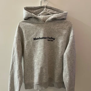 Grå hoodie Gina Tricot - Snygg grå hoodie från Gina Tricot med svart broderad text 'Manhattan Valley New York' på bröstet. Tröjan har huva, ribbade muddar och lång ärm. Perfekt för chill dagar och streetstyle. Mjuk och skön bomullsmix. Använd ett fåtal gånger💋