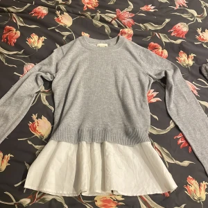 Grå tröja med vit volangkant - Supersöt grå tröja med lång ärm och en vit volangkant nedtill som ger en lager-på-lager look. Tröjan är ribbad vid midjan och har en klassisk rund halsringning. Perfekt för dig som gillar en stilren men ändå lekfull stil.