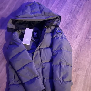 Grå dunjacka från Canada Goose - Säljer en grå dunjacka från Canada Goose med huva och klassisk patch på ärmen. Jackan är vadderad och har dragkedja framtill samt tryckknappar. Perfekt för kalla dagar och riktigt snygg streetstil.
