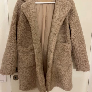 Beige teddyjacka från Kids Only - Mysig beige teddyjacka från Kids Only med stora framfickor och klassisk krage. Jackan är långärmad så den värmer hela kroppen, perfekt för kalla dagar. Tillverkad i mjukt teddy-material som ger en cool och avslappnad vibe.