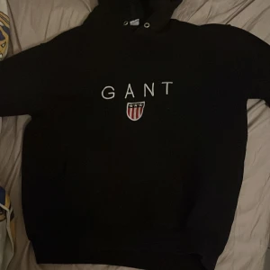 Svart hoodie från GANT med logga - Svart hoodie från GANT i storlek S med vit broderad logga och emblem på bröstet. Klassisk huva med snörning och ribbade muddar vid ärmslut och nederkant. Perfekt för chill dagar och enkel att matcha med jeans eller joggers.