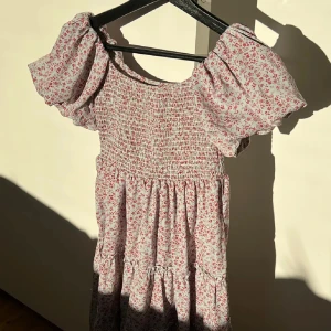 Vit blommig kortklänning med puffärm - Supersöt kortklänning i vitt med små röda blommor och puffiga kortärmar. Smockad överdel och volangig kjol ger en romantisk vibe. Perfekt för varma dagar och har en luftig passform. Materialet känns lätt och bekvämt.