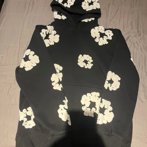 Svart hoodie från Fresh Team med blommor - Svart hoodie från Fresh Team med stora vita blommiga mönster över hela tröjan. Klassisk huva och magficka framtill. Skön och avslappnad passform, perfekt för dig som gillar streetwear och vill sticka ut lite extra.