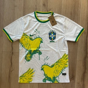 Vit Brasil Nike fotbollströja M - Snygg vit Brasilien fotbollströja från Nike med grönt och gult på krage och ärmslut. Framsidan har stora gula fåglar och Brasil-logga med broderade detaljer. Materialet är lätt och andas, perfekt för match eller träning.