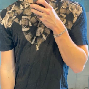 Marcelo Burlon t-shirt - Svart t-shirt från Marcelo Burlon med ett fett ormtryck runt halsen i gråa och beige toner. Klassisk passform och korta ärmar. Extremt coola slitningar vid nacken som du kan se på bild 4 och 5. Först till kvarn! 179 på bilden väger 67.
