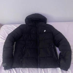 Svart pufferjacka Superdry S - Svart pufferjacka från Superdry i storlek s med huva och dragkedja. Jackan har två snedställda fickor med dragkedja, en innerficka på vänster sida, broderad logga på bröstet och diskret Superdry-tryck på ryggen. Fodrad insida och sportig look, perfekt för kalla dagar. Jackan har använts i en säsong. Den har inga skador och ser så gott som ny ut.