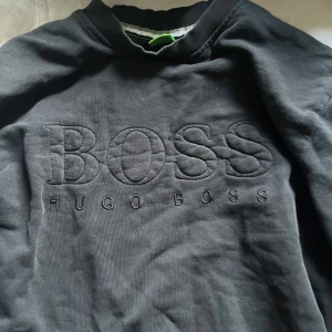 Svart sweatshirt från Hugo Boss - Svart sweatshirt från Hugo Boss med broderad logga framtill. Tröjan har rund halsringning med ribbad kant och långa ärmar. Tillverkad i mjuk bomull, perfekt för en clean och stilren look.