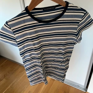 Randig topp från Brandy Melville - Säljer en snygg randig topp från Brandy Melville i blå och vitt. T-shirten har rund hals och korta ärmar, perfekt för en chill och stilren look. Materialet är mjuk bomull som känns skönt mot huden. Passar dig som gillar klassiska ränder och enkel design.