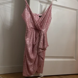 Rosa glittrig klänning Double Crazy - Rosa paljettklänning från Double Crazy med omlott-v-ringning, smala axelband och draperad detalj framtill. Klänningen har en figurnära passform och dragkedja bak. Perfekt för dig som vill sticka ut med extra glitter och glam.