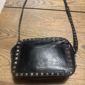 Svart axelväska Valentino med nitar - Cool svart axelväska från Valentino i äkta skinn med silverfärgade nitar runt kanterna och på axelremmen. Väskan har en rektangulär form, justerbar rem och dragkedja upptill. Perfekt storlek för det viktigaste och ger en edgy vibe till din outfit. Har få defekter därför de låga priset.