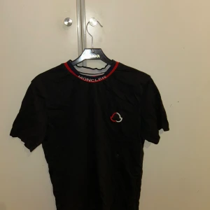 Svart Moncler t-shirt med logga - Svart t-shirt från Moncler med korta ärmar och rund halsringning med röd och blå kant. Moncler-logga broderad på bröstet och text på kragen. Skön passform och stilren design, perfekt för en trendig look.