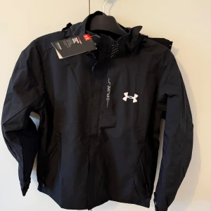 Svart vindjacka från Under Armour - Snygg svart vindjacka från Under Armour med huva och dragkedja framtill. Jackan har en vit logga på bröstet och är perfekt för blåsiga dagar. Lätt och smidig design som funkar till många outfits. Storlek M men passar S också,Aldrig använd