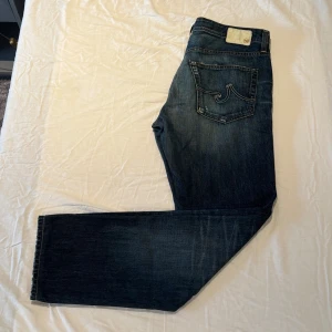 Jeans från Adriano Goldschmied - Snygga mörkblå jeans från AG (Adriano Goldschmied) med designade slitningar framtill. Jeansen har raka ben och normal/slim passform. Adriano Goldschied är ett lyxmärke som säljer jeans för mellan 2500-5000kr. Skicket på jeansen är fantastisk, inga tecken på användning. Storlek 34/34.