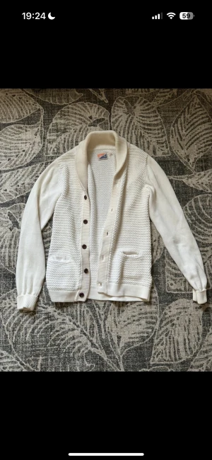 Vit kofta med bruna knappar - Snygg vit kofta från Campus med grovstickad framsida och slätstickade ärmar. Koftan har bruna knappar, två fickor framtill och en bred krage som ger en chill vibe. Perfekt för lager-på-lager och passar till jeans eller kjol.