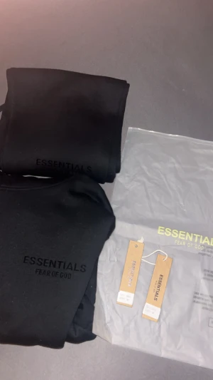 Svart  Essentials Fear of God tracksuit - Svart hoodie från Essentials Fear of God med stilren logga på bröstet. Tröjan har huva, känguruficka och ribbade muddar. Materialet är mjukt och skönt, perfekt för chill dagar. Passformen är normal och ärmarna är långa. Snygg och enkel design som funkar till streetwear.