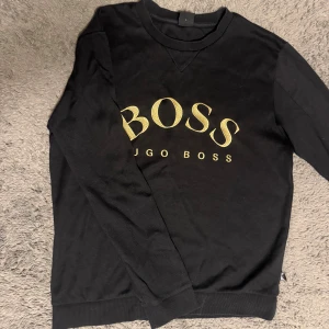 Svart sweatshirt från Hugo Boss - Svart sweatshirt från Hugo Boss med stort BOSS-tryck i guld på bröstet. Tröjan har rund hals, ribbade muddar och lång ärm. Perfekt för dig som gillar stilrena och lyxiga märkesplagg med tydlig logga.