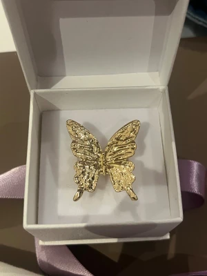 Guldfärgad ring med fjäril - Unik guldfärgad ring med en stor, detaljerad fjäril som dekoration från glitter. Fjärilen har en glansig och texturerad yta som ger ett coolt statement på handen. Perfekt för dig som vill sticka ut med din stil och gillar djurmotiv. Ringen levereras i en fin presentask med lila band.