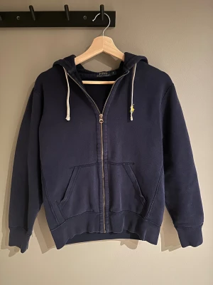 Marinblå hoodie från Polo Ralph Lauren - Snygg marinblå hoodie från Polo Ralph Lauren i storlek S. Tröjan har dragkedja, huva med vita snören och klassisk gul broderad logga på bröstet. Mjuk bomullskvalitet och två fickor framtill. Perfekt för chill dagar och streetstyle.