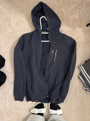 Mörkblå hoodiejacka Sail Racing - Mörkblå hoodiejacka från Sail Racing med dragkedja och snörning i huvan. Jackan har en bröstficka med dragkedja och logotyp på framsidan. Perfekt för chill dagar och enkel att matcha med jeans eller joggers.