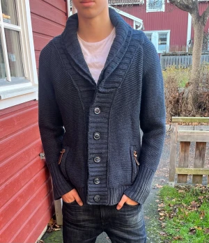 Mörkblå stickad cardigan från Superdry - Säljer en mörkblå stickad cardigan från Superdry med grov struktur, bred krage och snygga ribbade muddar. Koftan har knappar framtill, två fickor med dragkedja och läderdetaljer. Perfekt för kyliga dagar och riktigt skön att ha på sig.